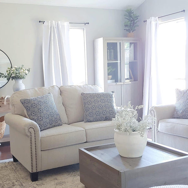 Beige fabric sofa with rolled arms, detailed nailhead trim, and dark wooden feet, styled in a bright living room.