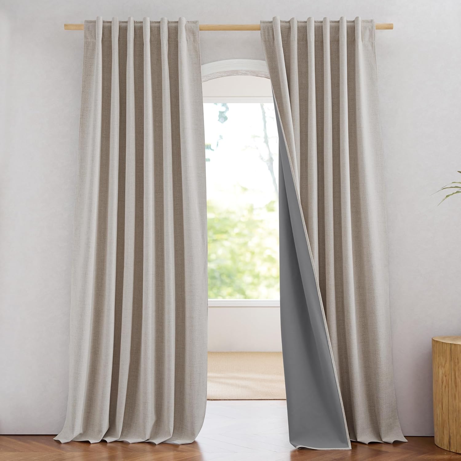 Beige linen back tab blackout curtains with dark grey lining, hanging on a wooden rod over an arched window.