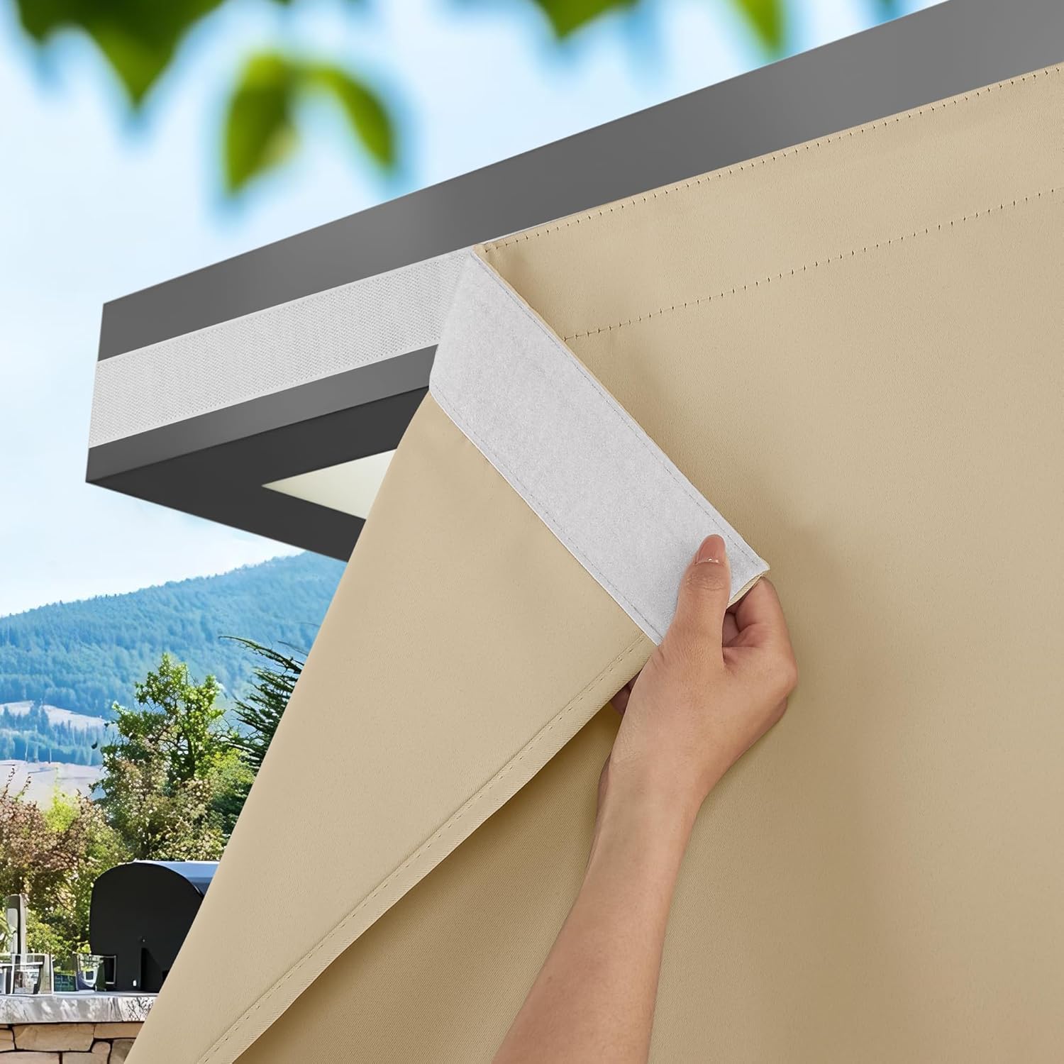Beige outdoor privacy curtain attaches to dark pergola frame with hook-and-loop fastener. Scenic patio background.