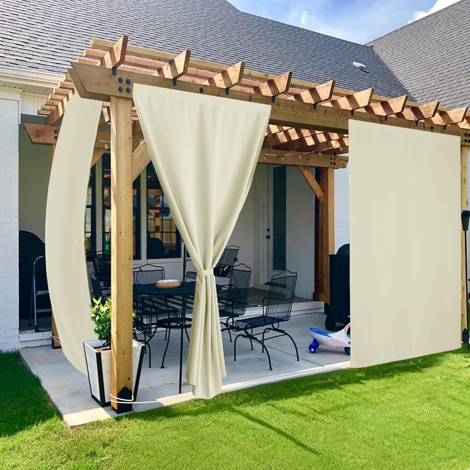 Beige outdoor privacy curtains hanging from a wooden pergola over a patio, one panel tied back.