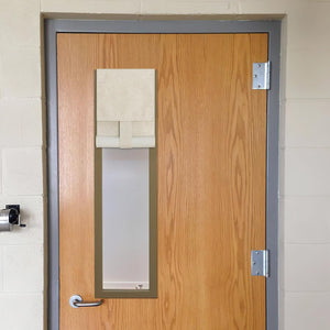 Beige fabric roll-up privacy shade covering a narrow glass window on a wooden classroom door.