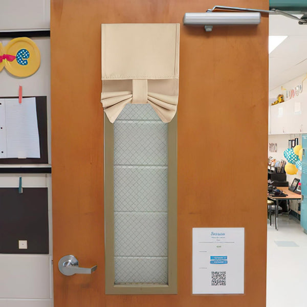 Beige fabric tie-up door window shade covering the upper portion of a classroom door window for privacy.