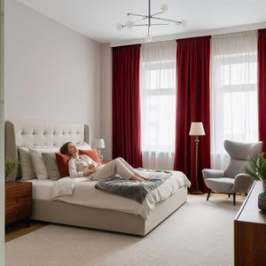 Elegant bedroom with a beige tufted wingback bed, deep red and sheer white curtains. A person relaxes on the bed.