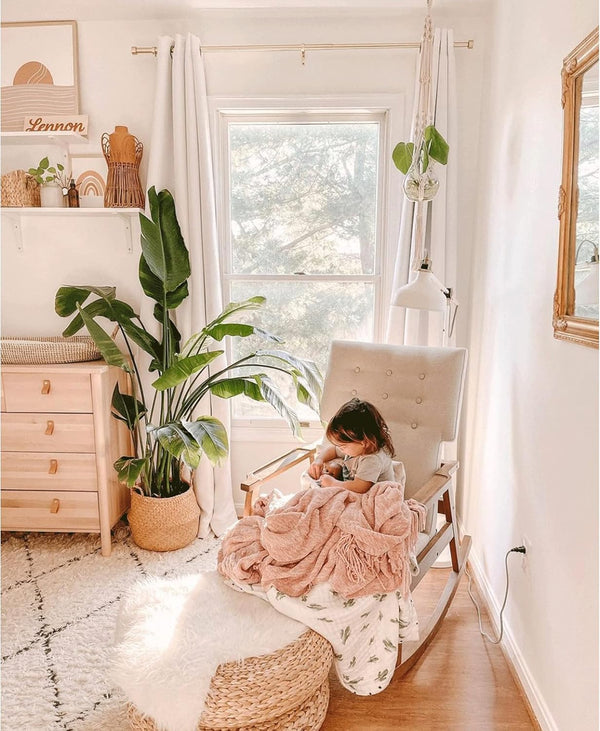 Child in cozy neutral upholstered rocking chair with pink knit blanket and woven pouf.