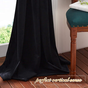 Black velvet blackout curtain hanging full length by a window, pooling on wooden floor, with green trees visible outside.