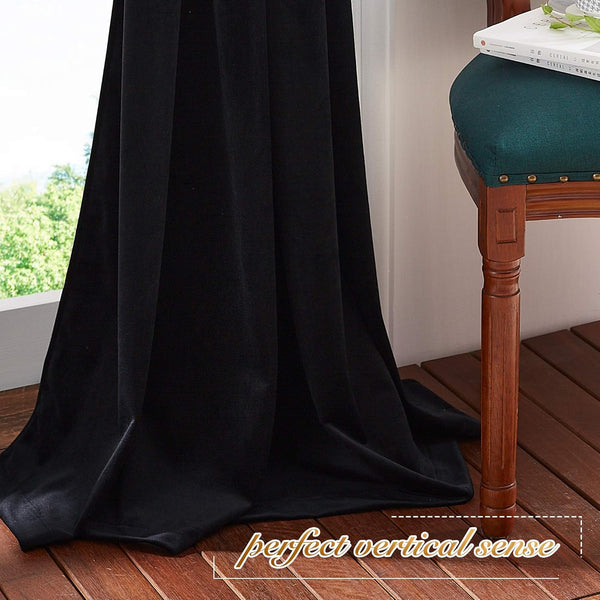Black velvet blackout curtain hanging full length by a window, pooling on wooden floor, with green trees visible outside.