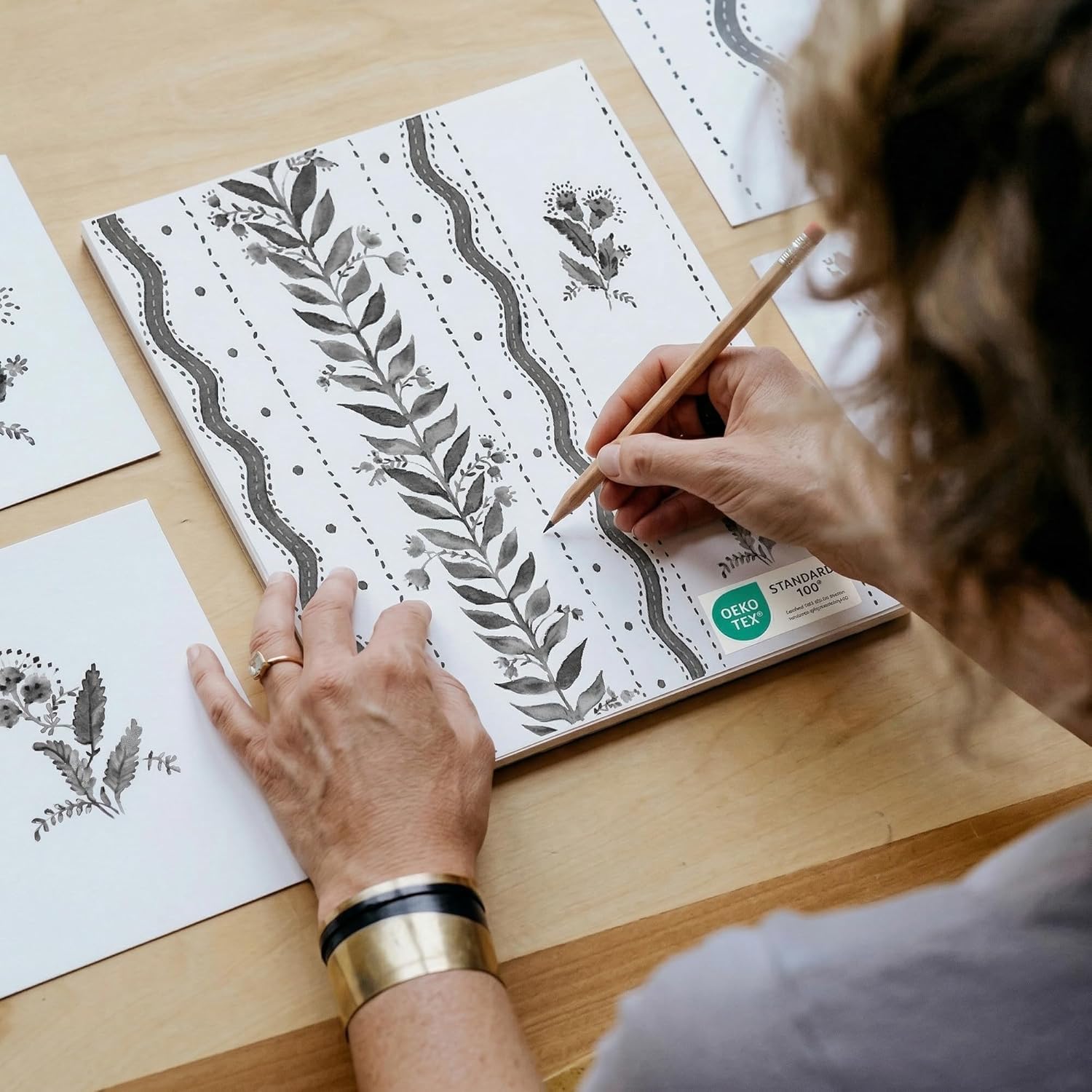 Hands drawing a black and white floral pattern with leafy vines. Paper features an OEKO-TEX label.
