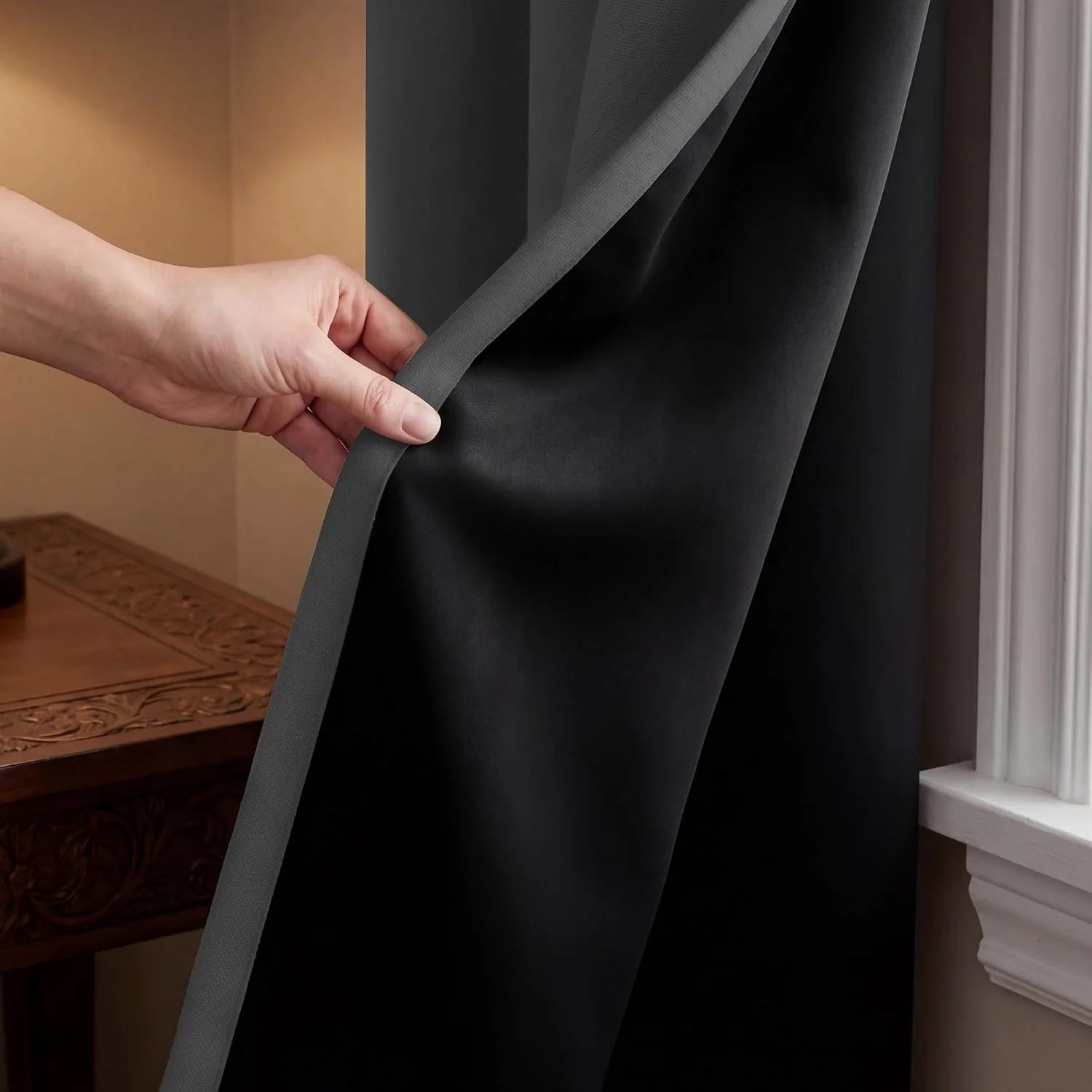 Hand pulling back a dark blackout curtain to show its textured grey lining by a window.