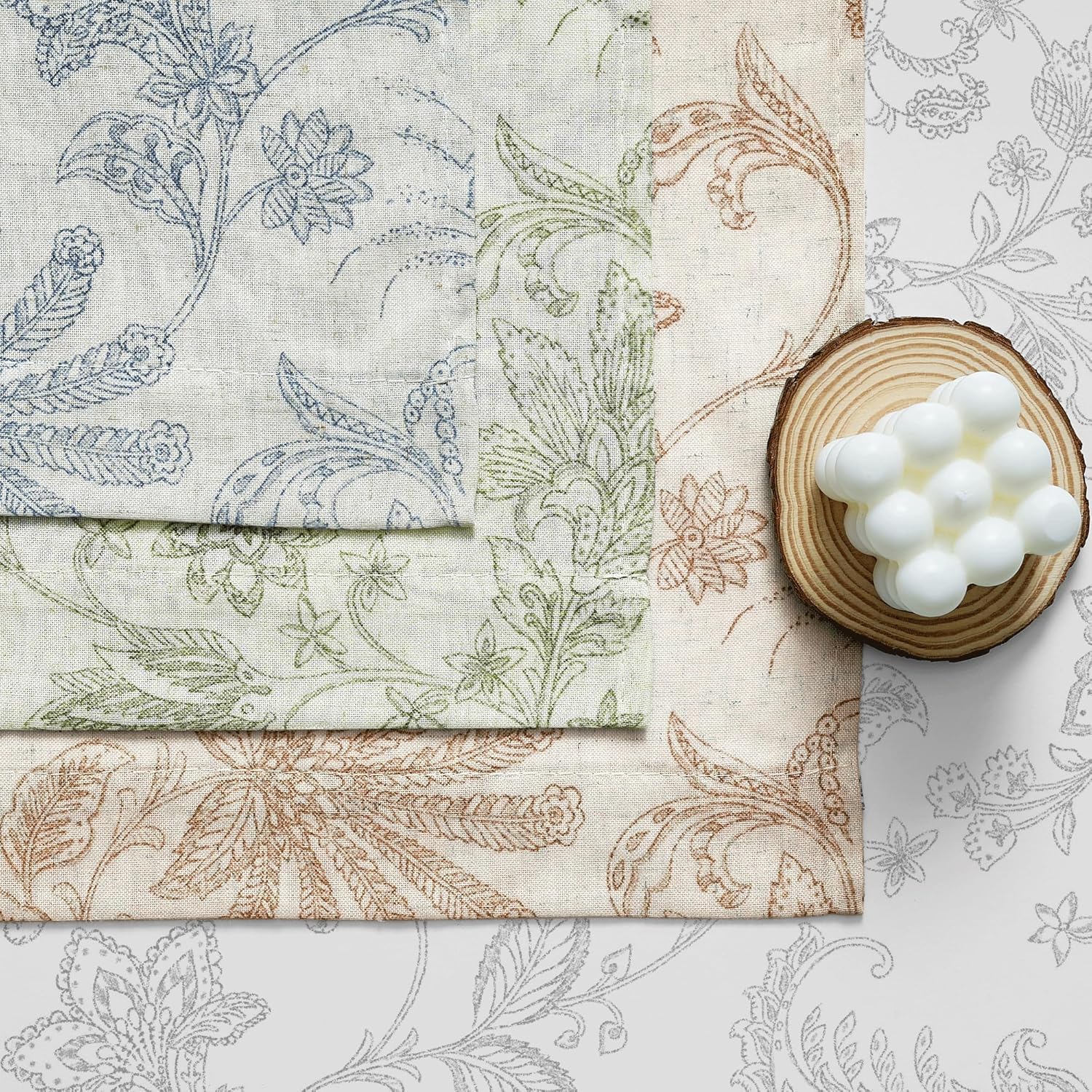 Folded linen fabric swatches feature blue, green, and brown floral patterns, styled with a white bubble candle.