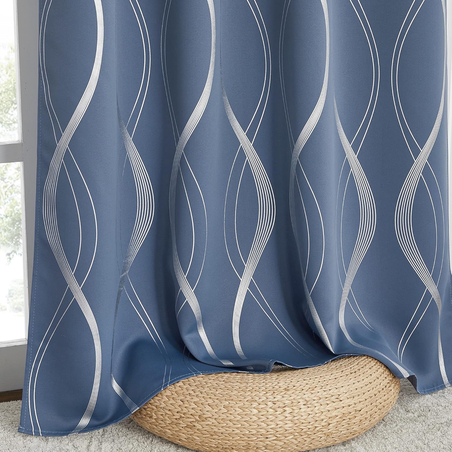 Blue blackout curtain panel with metallic silver geometric wave pattern, pooling on a woven pouf by a window.