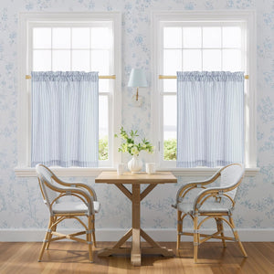 Pair of blue and white striped cafe curtains with a rod pocket top, hanging on a gold rod in a bright kitchen nook.