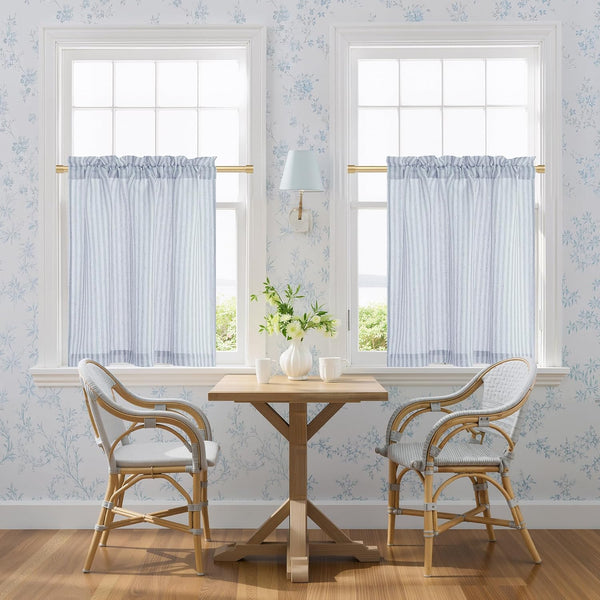 Pair of blue and white striped cafe curtains with a rod pocket top, hanging on a gold rod in a bright kitchen nook.
