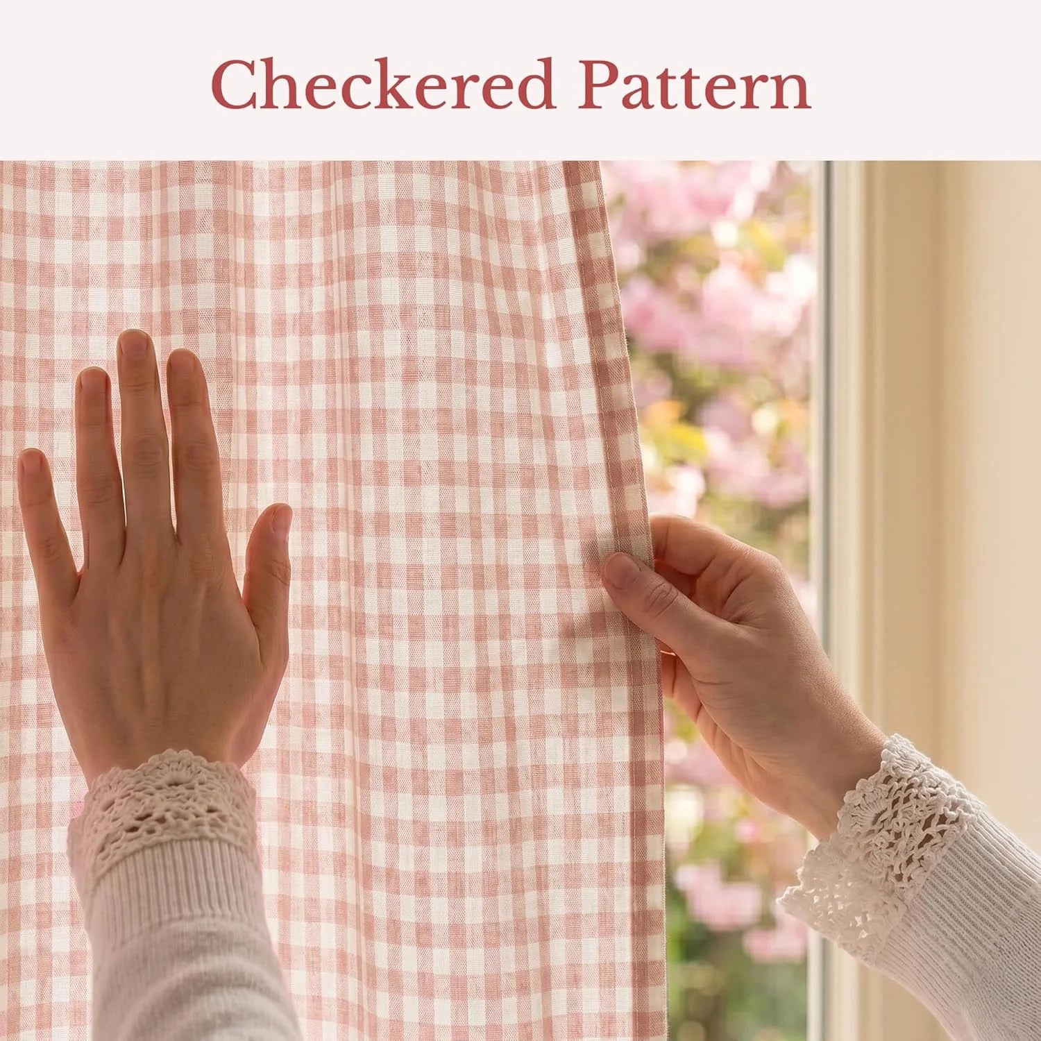 A hand opens blush pink and white gingham checkered curtains by a window with blooming pink flowers outside.