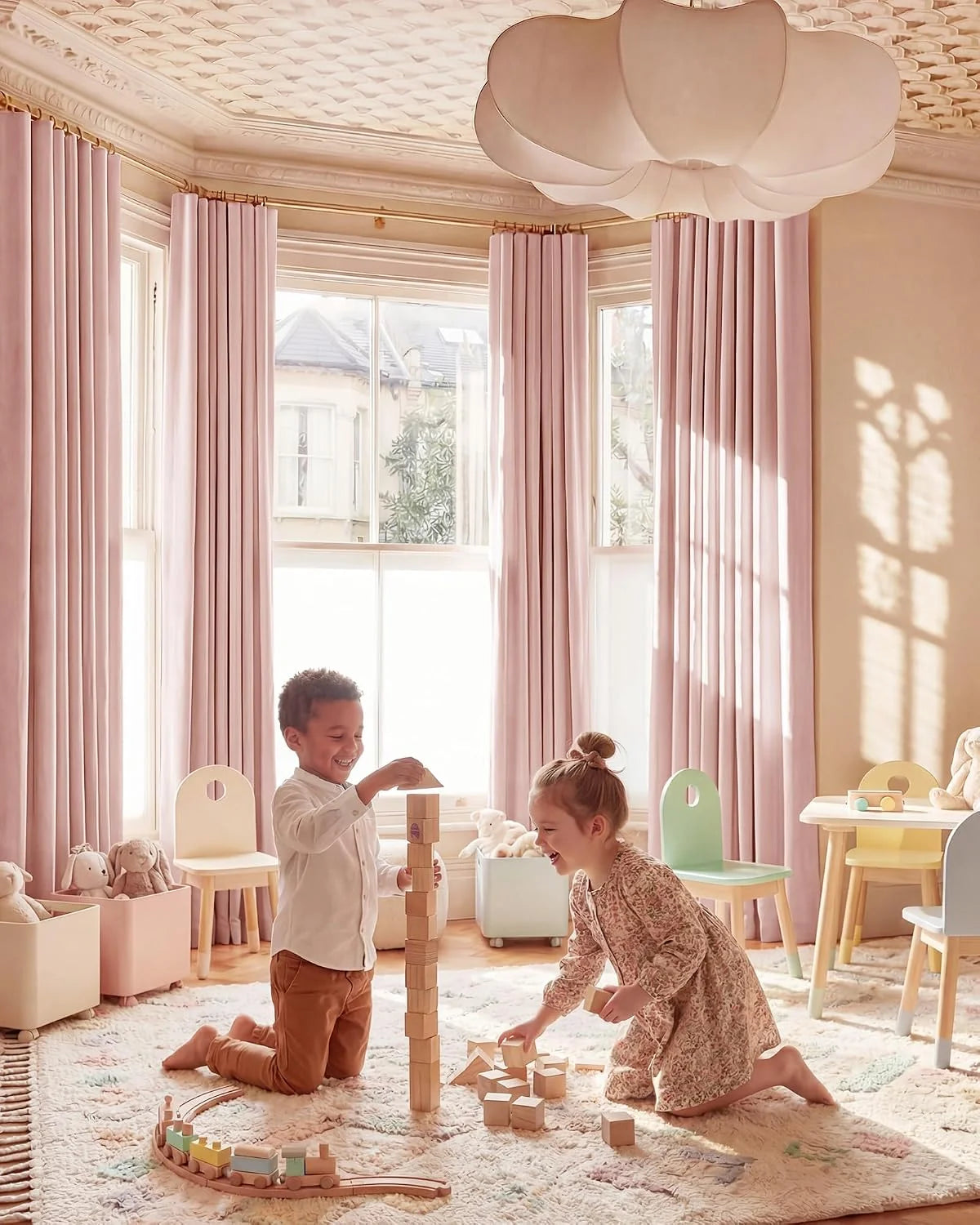 Two children playing with wooden blocks in a sunlit playroom with blush pink curtains