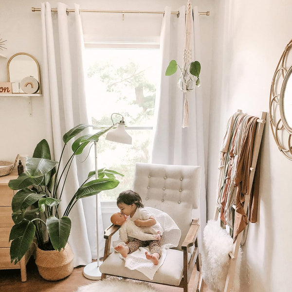 Bright boho nursery featuring white grommet curtains, cream tufted armchair, and a large plant in a woven basket.