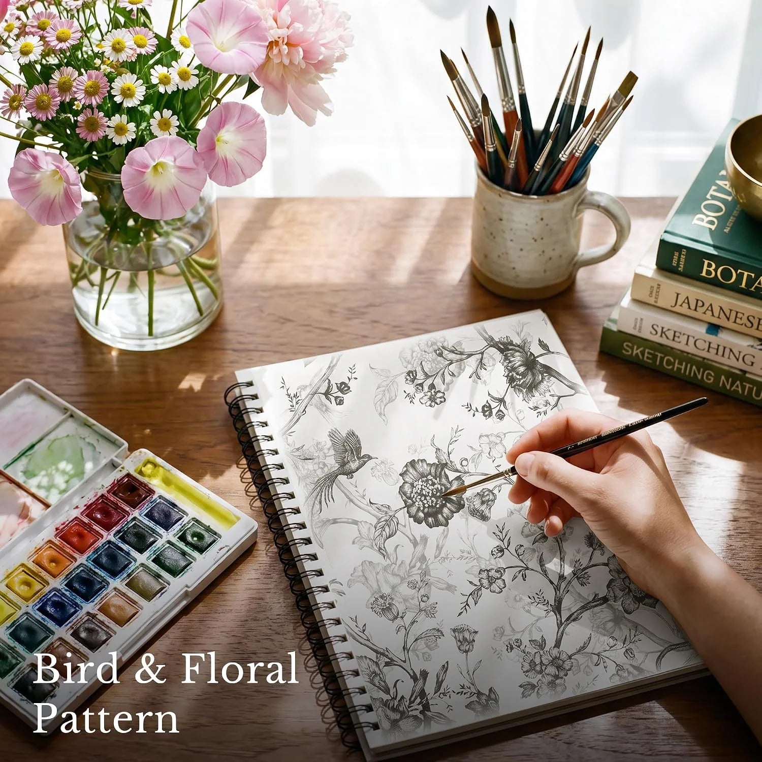 Hand painting a bird and floral pattern in a sketchbook beside watercolors and flowers