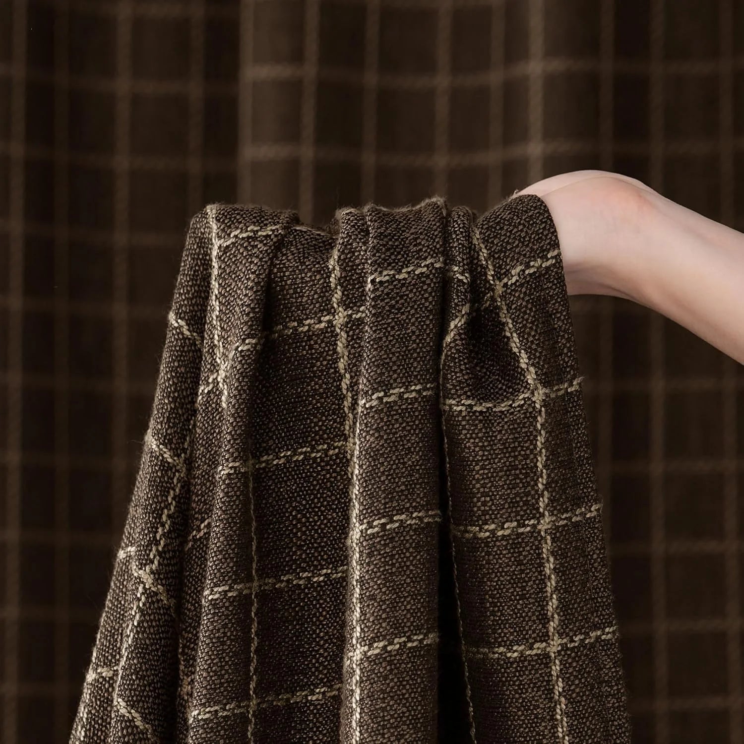 Hand holding folded dark brown woven fabric with light textured plaid pattern, matching background.