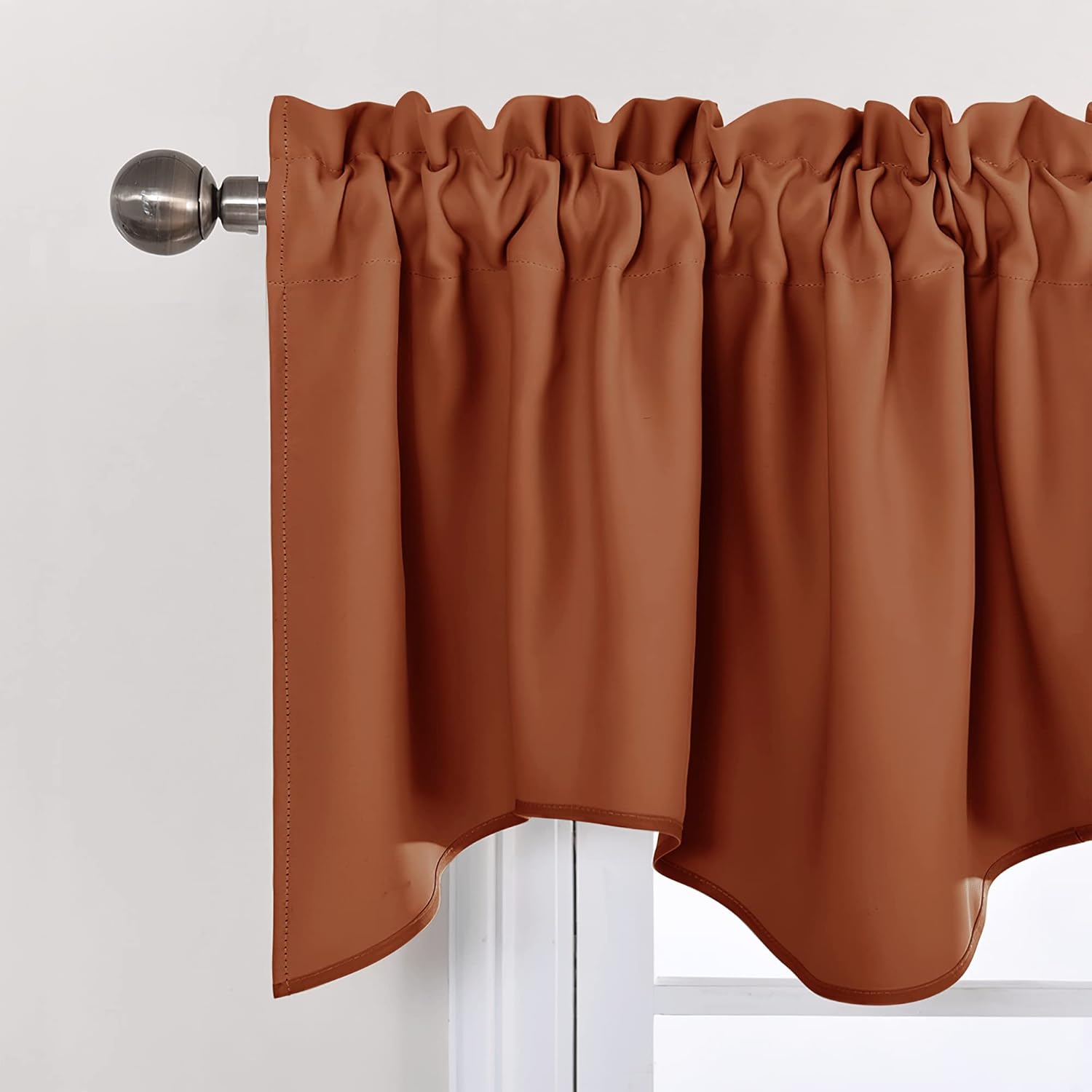 Burnt orange rod pocket valance with a scalloped bottom edge hanging on a bronze metal curtain rod over a window.