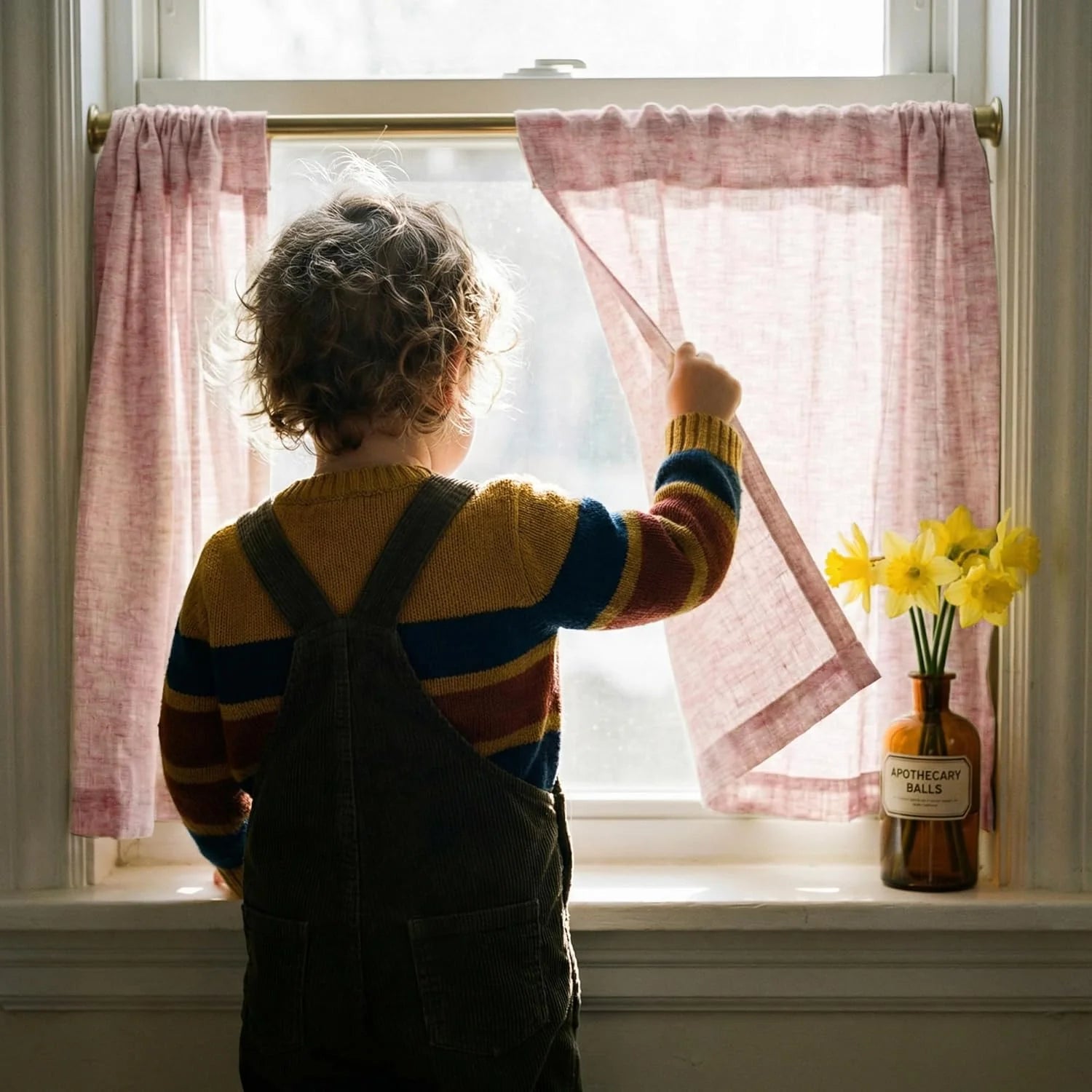 Curly-haired child holding back a pink sheer curtain, looking out a bright window with daffodils.