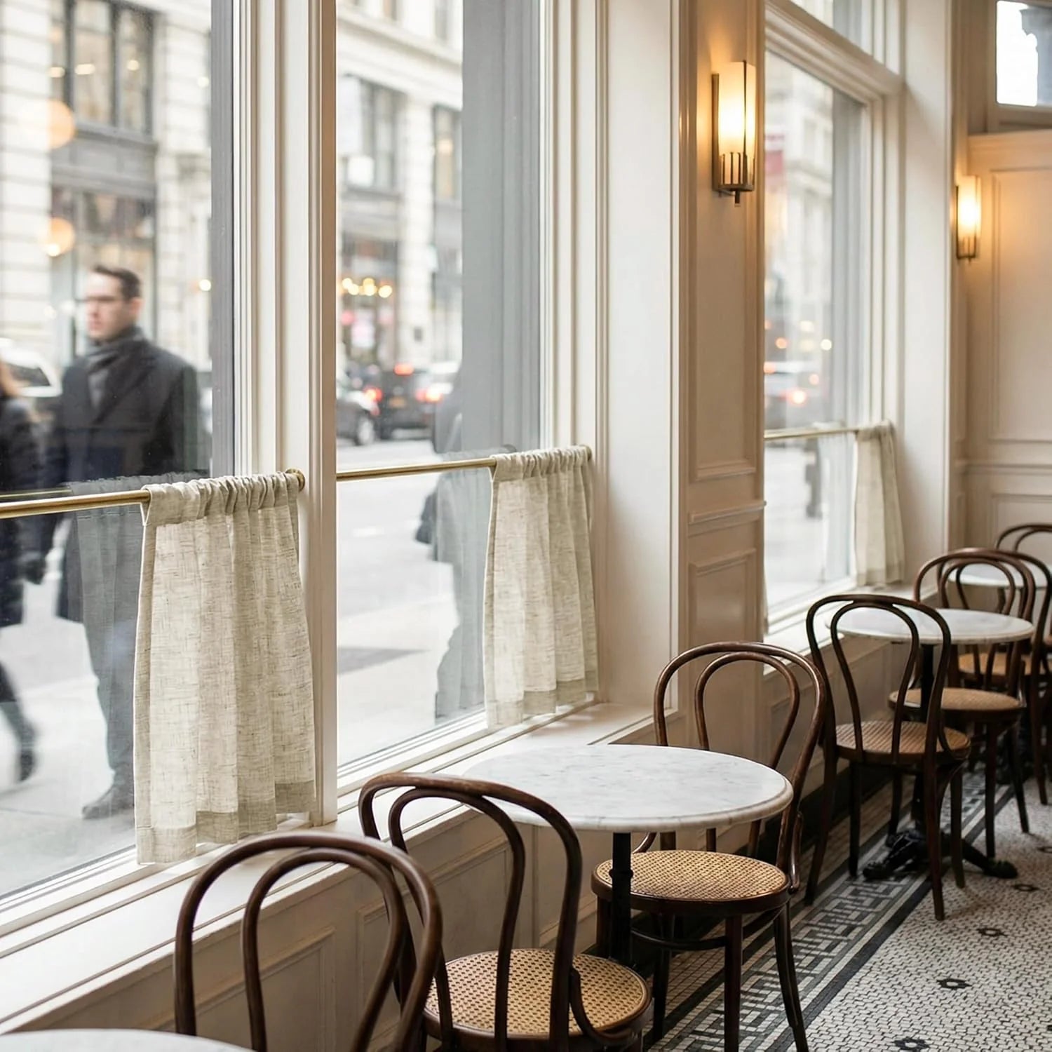 Classic cafe interior with marble tables, bentwood chairs, and sheer curtains beside a city street window.