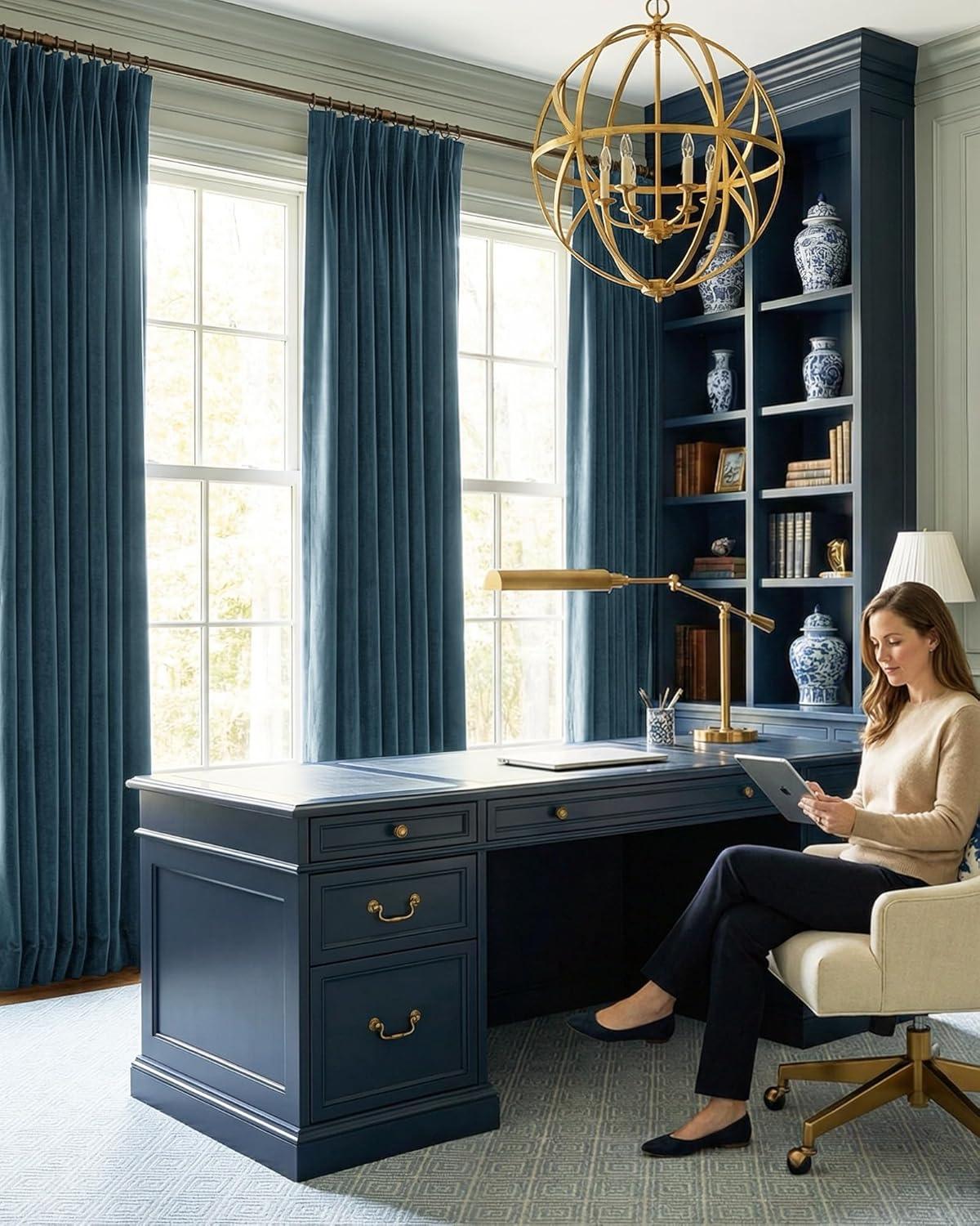 Elegant navy blue desk, matching built-in bookcase, and velvet curtains in a home office. Gold chandelier.