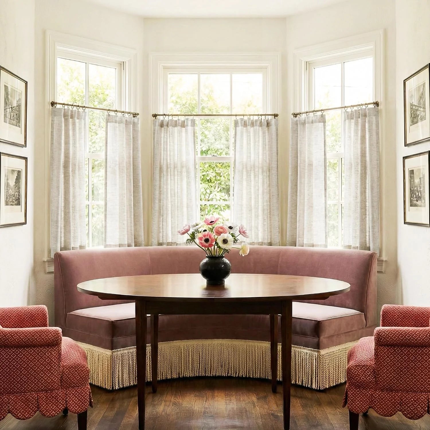 Cozy dining nook with pink velvet banquette, round wood table, two red chairs, and sheer curtains.