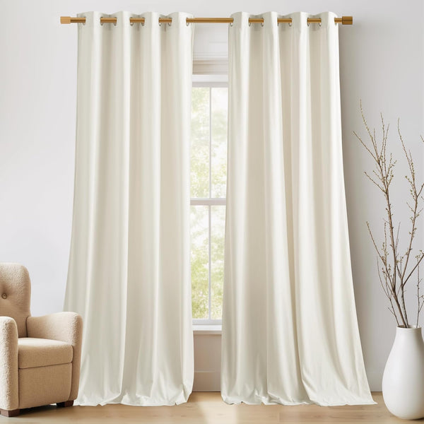 Pair of cream grommet top blackout curtains hanging on a wooden rod, pooling on the floor.