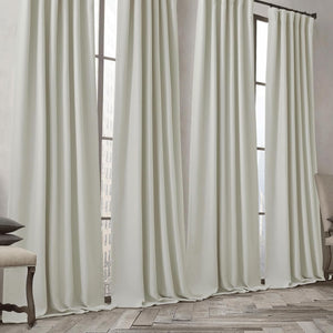 Pair of light cream tailored pinch pleat drapes hanging from a black rod, pooling slightly on a rustic wooden floor.