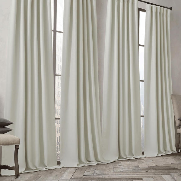 Pair of light cream tailored pinch pleat drapes hanging from a black rod, pooling slightly on a rustic wooden floor.