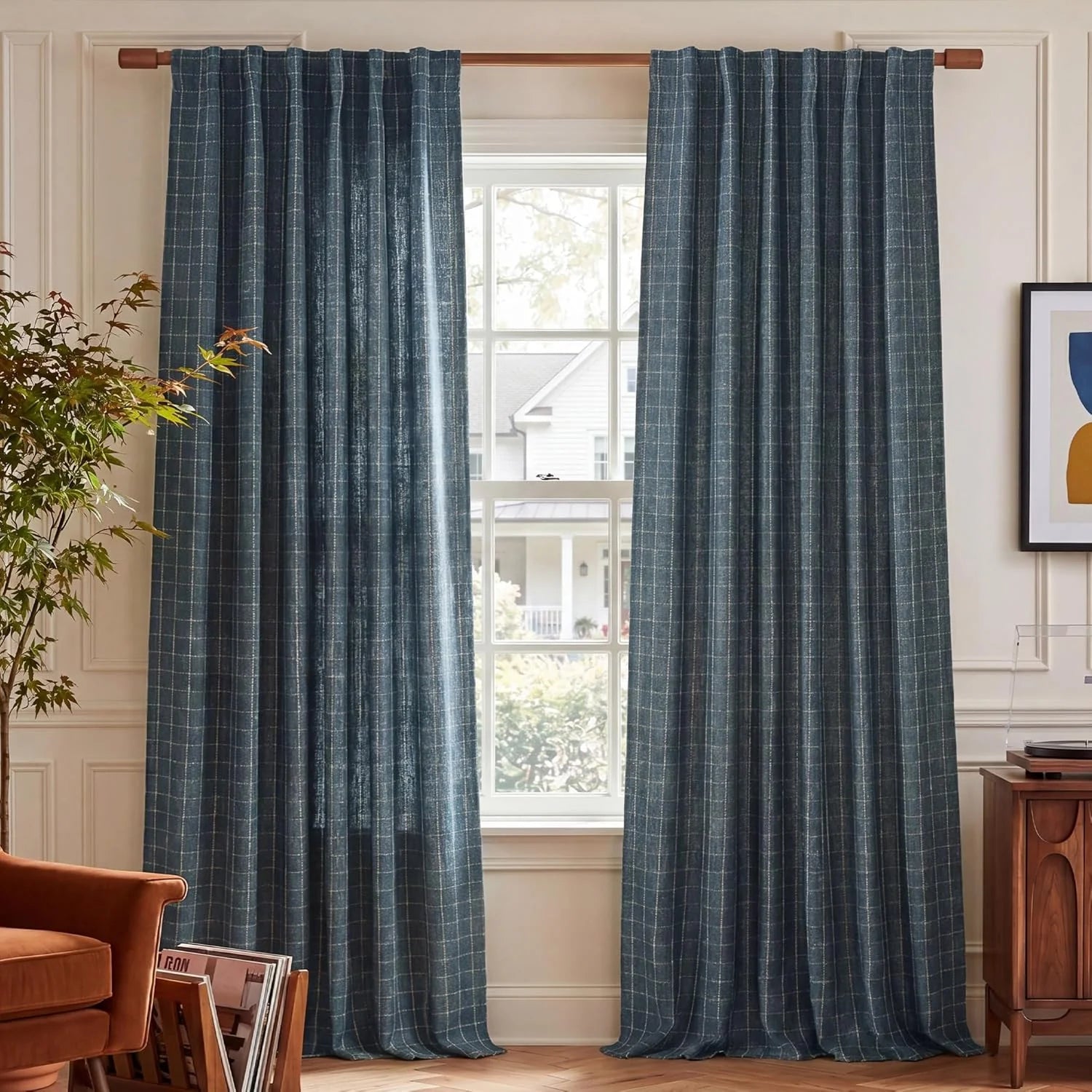 Pair of dark blue grid patterned curtains on a wooden rod in a sunlit living room with an armchair and plant.
