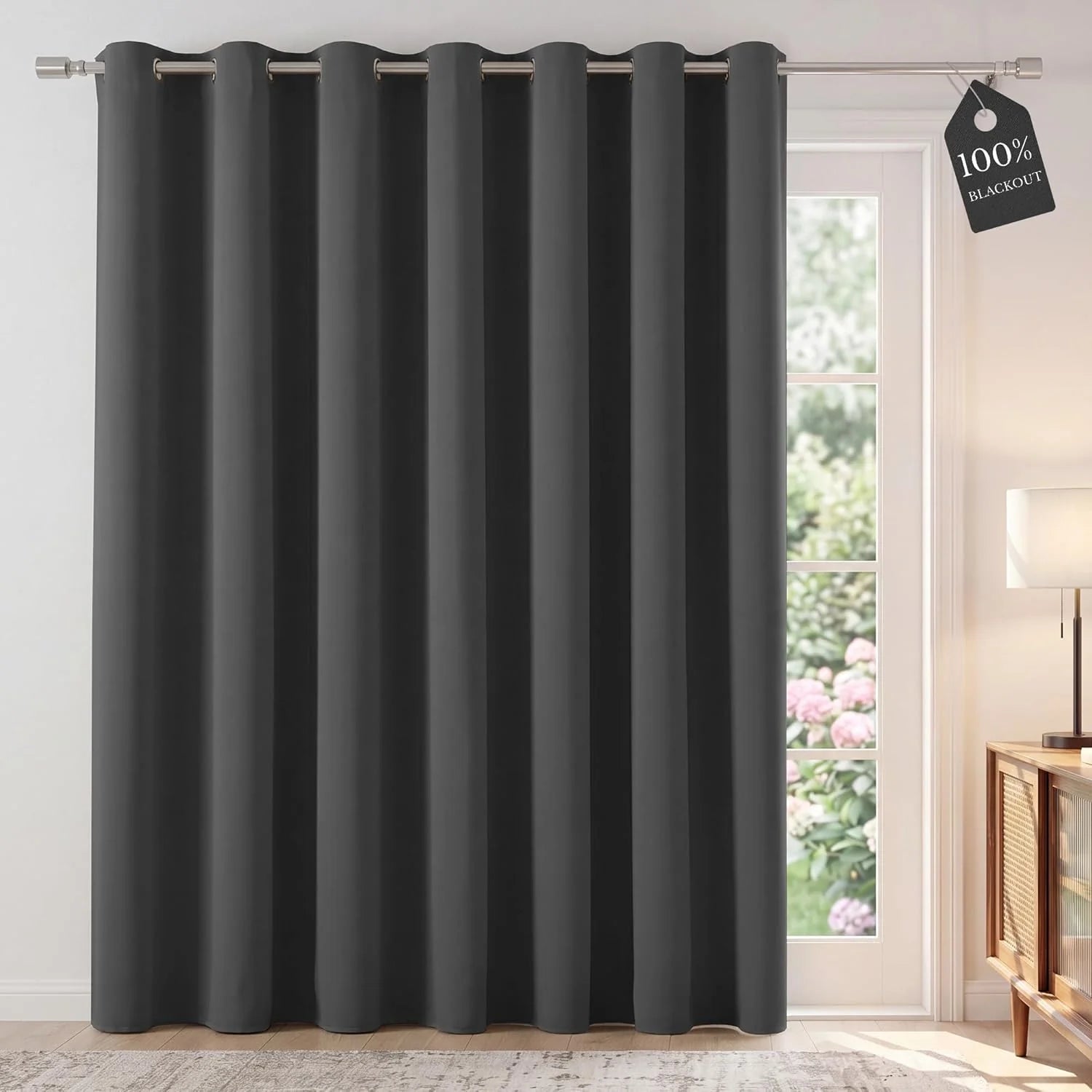 Dark gray blackout grommet curtain panel covering a glass patio door, with a '100% BLACKOUT' tag visible.