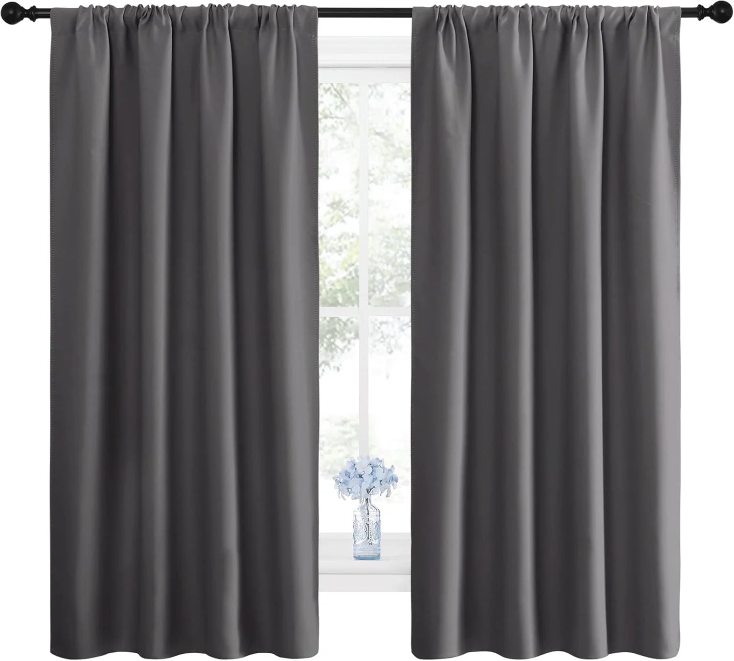 Pair of dark gray blackout curtains with a rod pocket header style, hanging over a bright window with a small vase of blue flowers.