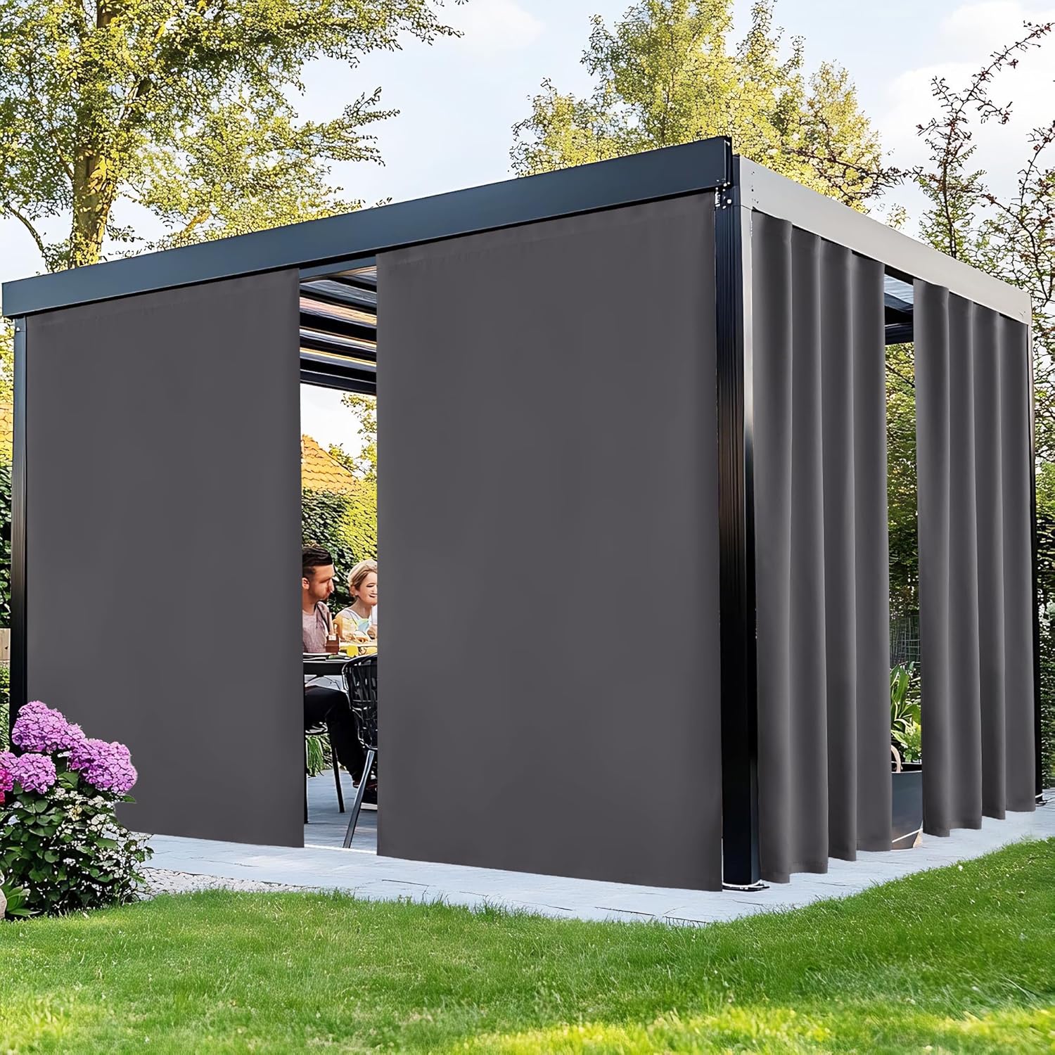 Dark gray privacy screens enclose a black modern pergola where a couple dines outdoors in a sunny garden.
