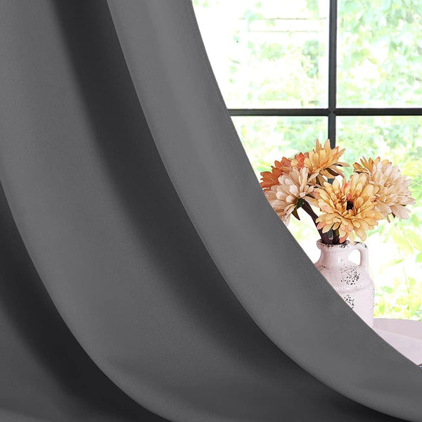 Close-up of dark grey smooth fabric curtain with deep folds. Window and vase with flowers in background.