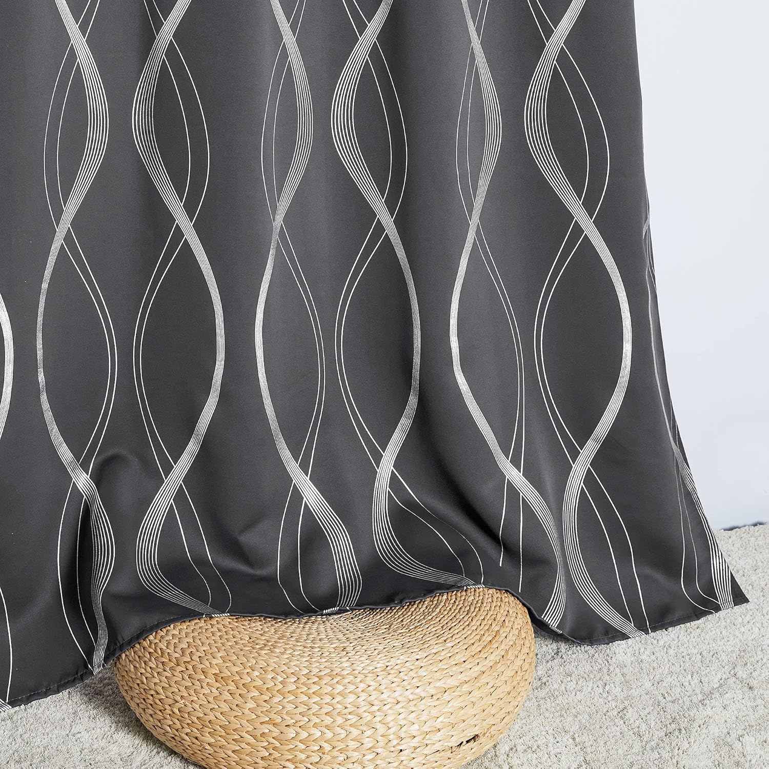 Dark grey curtains featuring wavy silver abstract lines, hanging and pooling on a wicker pouf and light rug.
