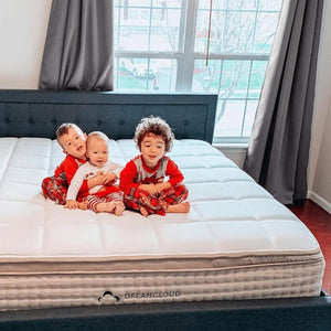 Three happy children sit on a white quilted Dreamcloud mattress with a dark tufted upholstered bed frame.