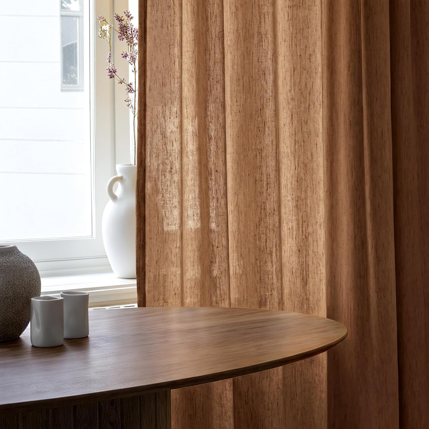 Earthy brown textured drapes with a subtle vertical weave, hanging by a bright window. A wooden table is in the foreground.