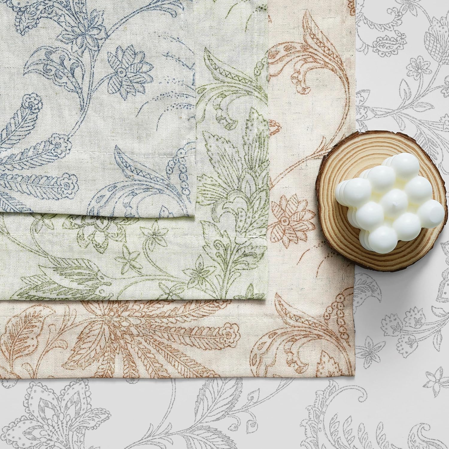 Stacked natural linen fabric swatches with blue, green, and rust floral patterns. White bubble candle on wood.