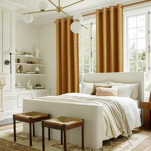 Warm golden brown grommet top curtains frame tall bedroom windows, beside an upholstered bed.