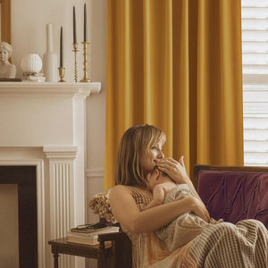 Golden yellow solid drapes brighten a window in a cozy living room, next to a woman holding a baby.