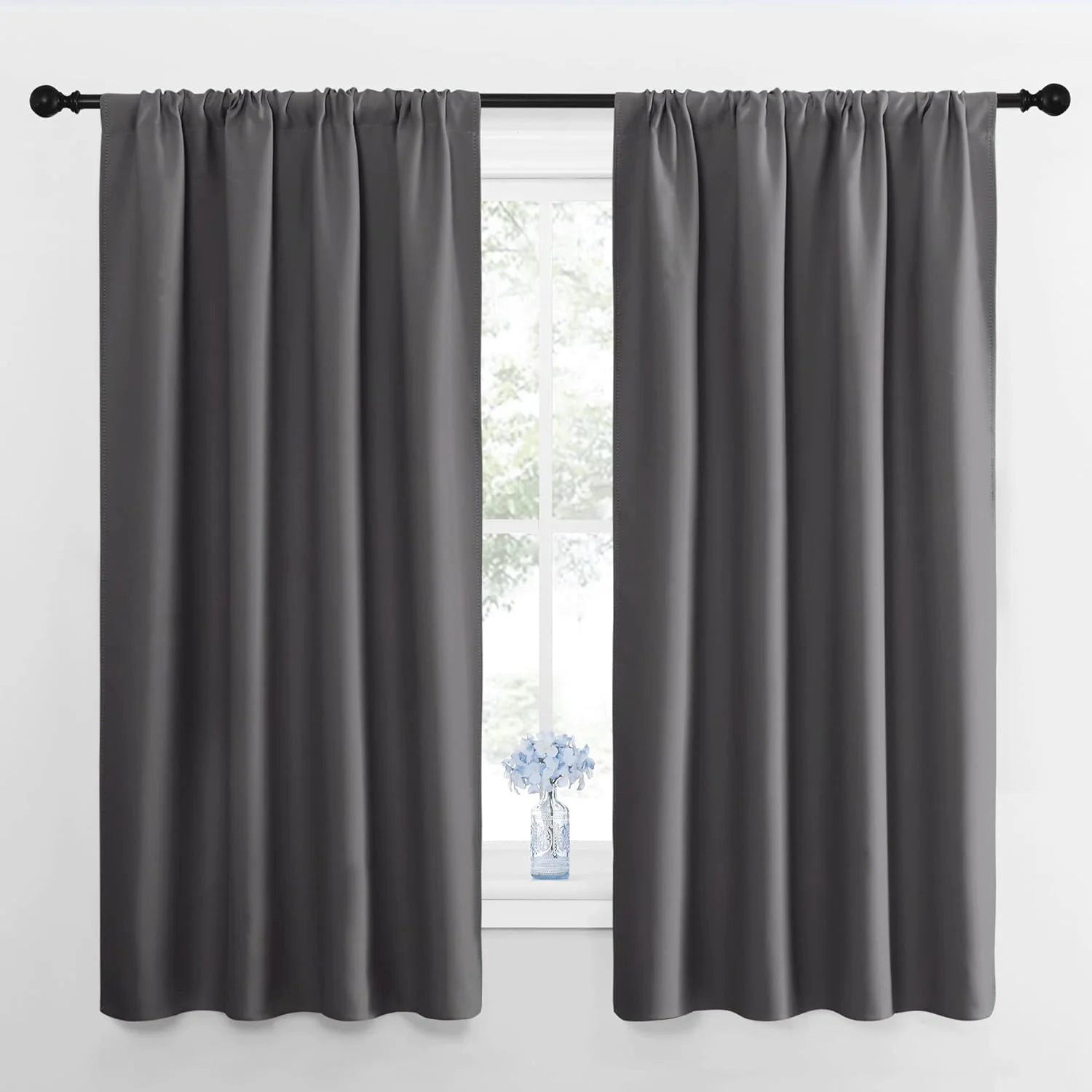 Pair of dark gray blackout curtains with a rod pocket top, hanging on a black rod over a window. A small blue vase is on the sill.
