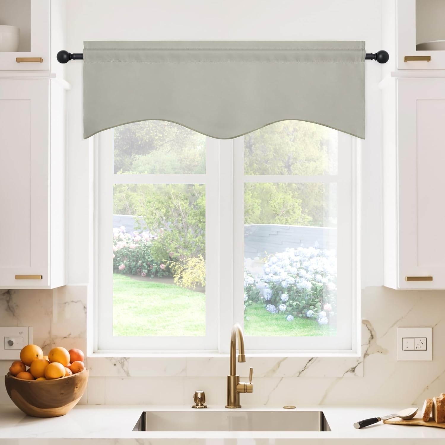 Light greige fabric window valance with scalloped bottom edge on black rod, above a kitchen sink.