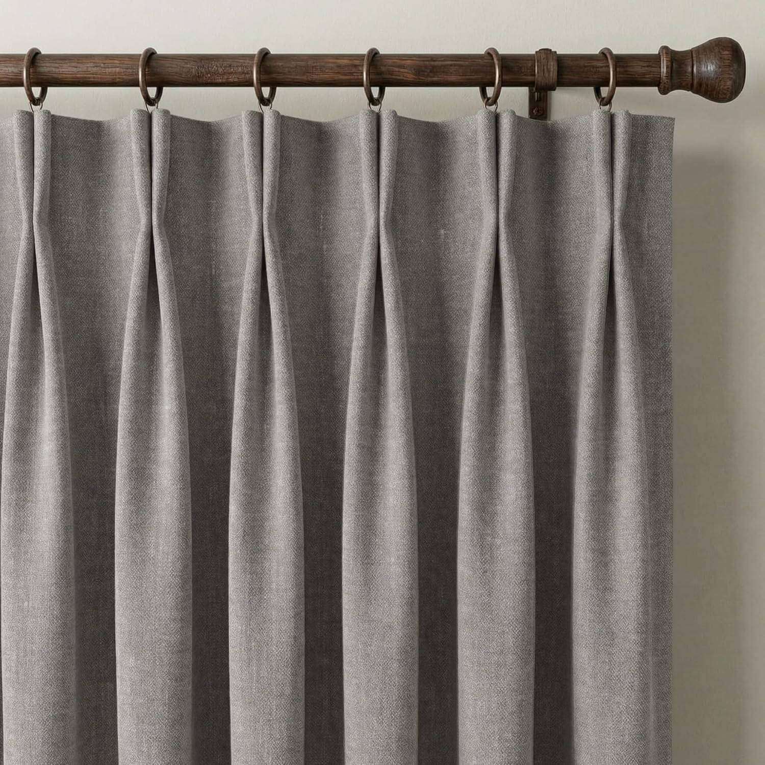 Close-up of grey textured pinch pleat curtains hanging from a dark wood rod with antique bronze rings.