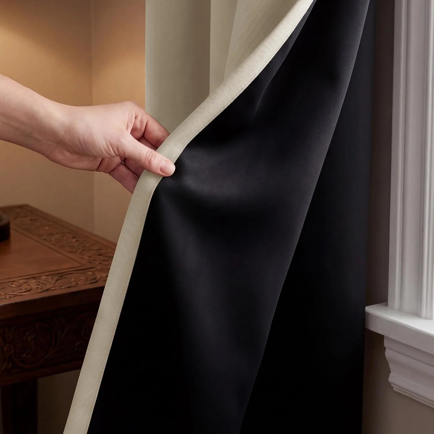 Hand pulls back a beige curtain, revealing its black blackout lining, next to a window and ornate wood table.
