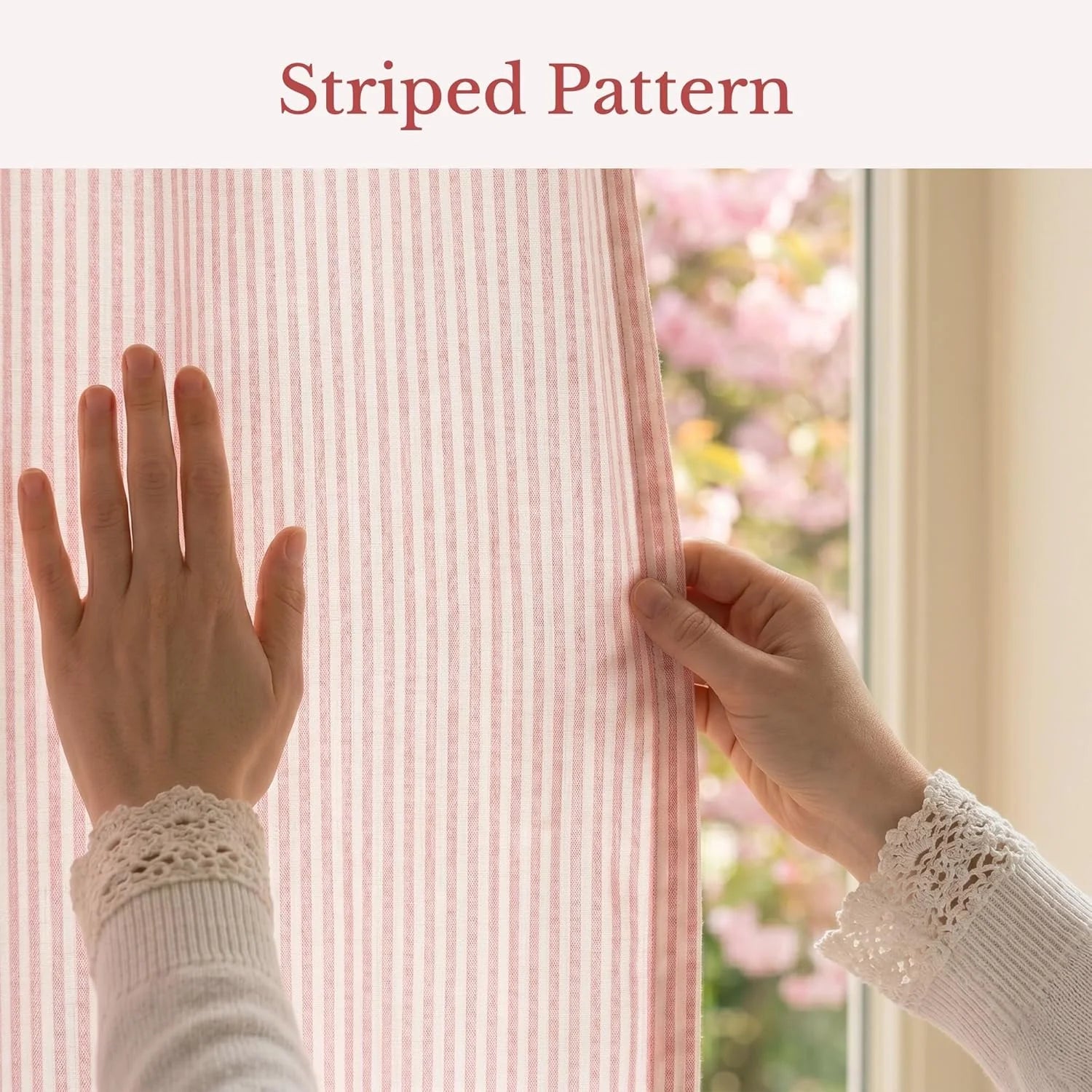 Hands gently touch a pink and white striped curtain fabric, by a sunlit window with blooming trees outside.