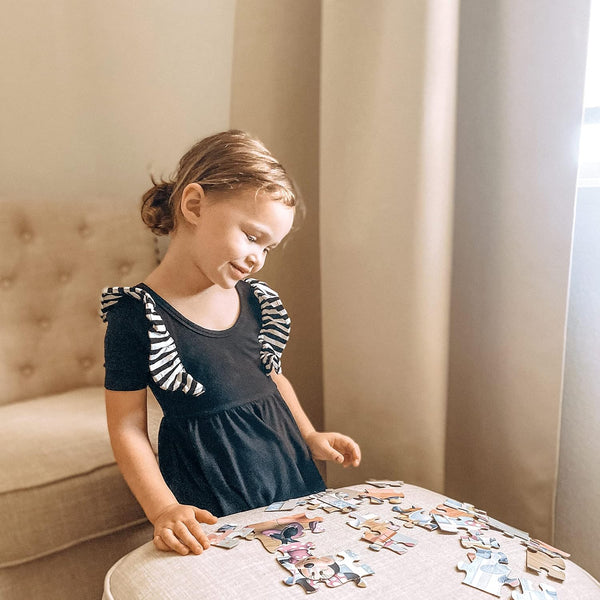 Young girl in black dress with striped sleeves playing a colorful cartoon character jigsaw puzzle on an ottoman.