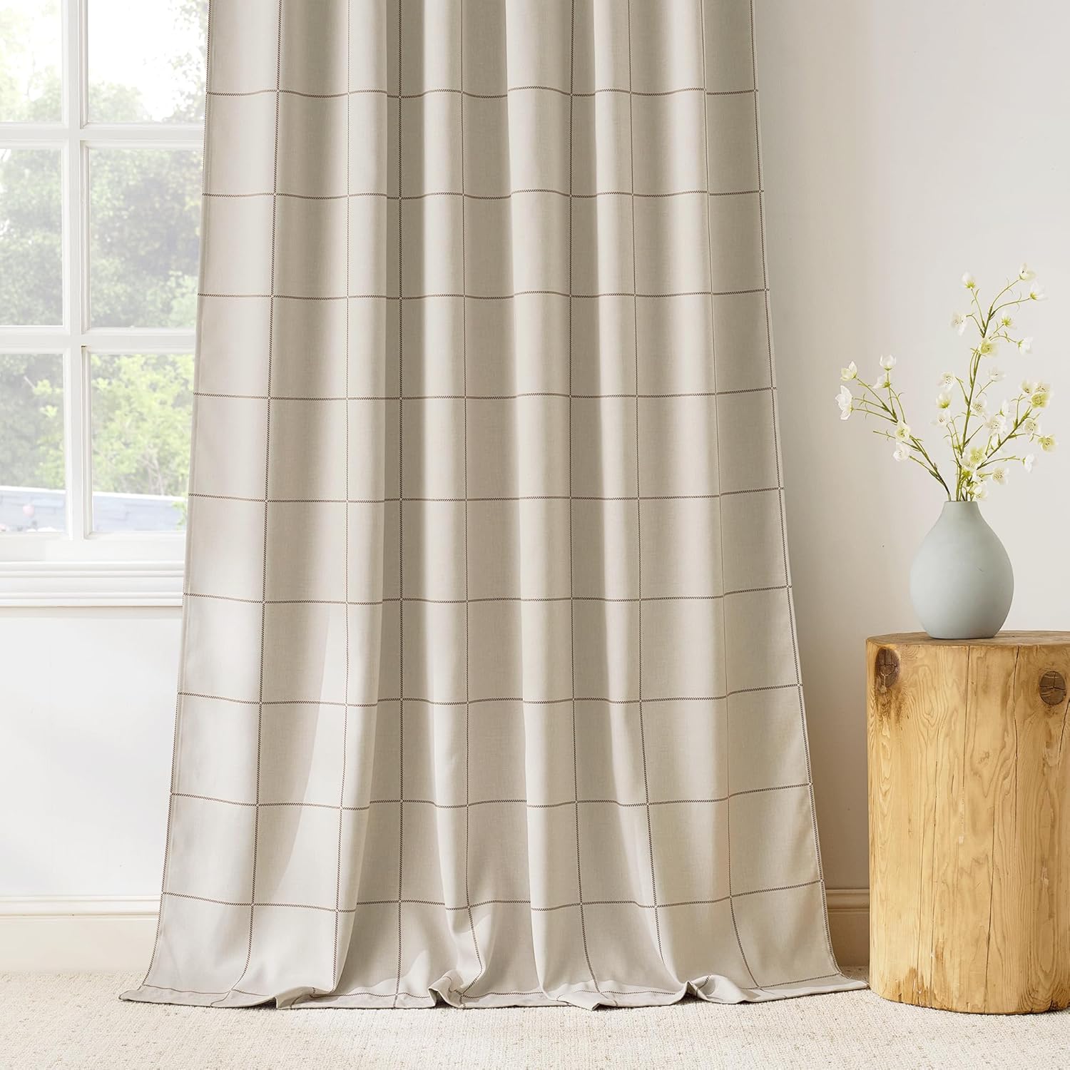 Light beige fabric curtain panel with subtle dark grid pattern hangs by a window, pooling on a beige carpet.