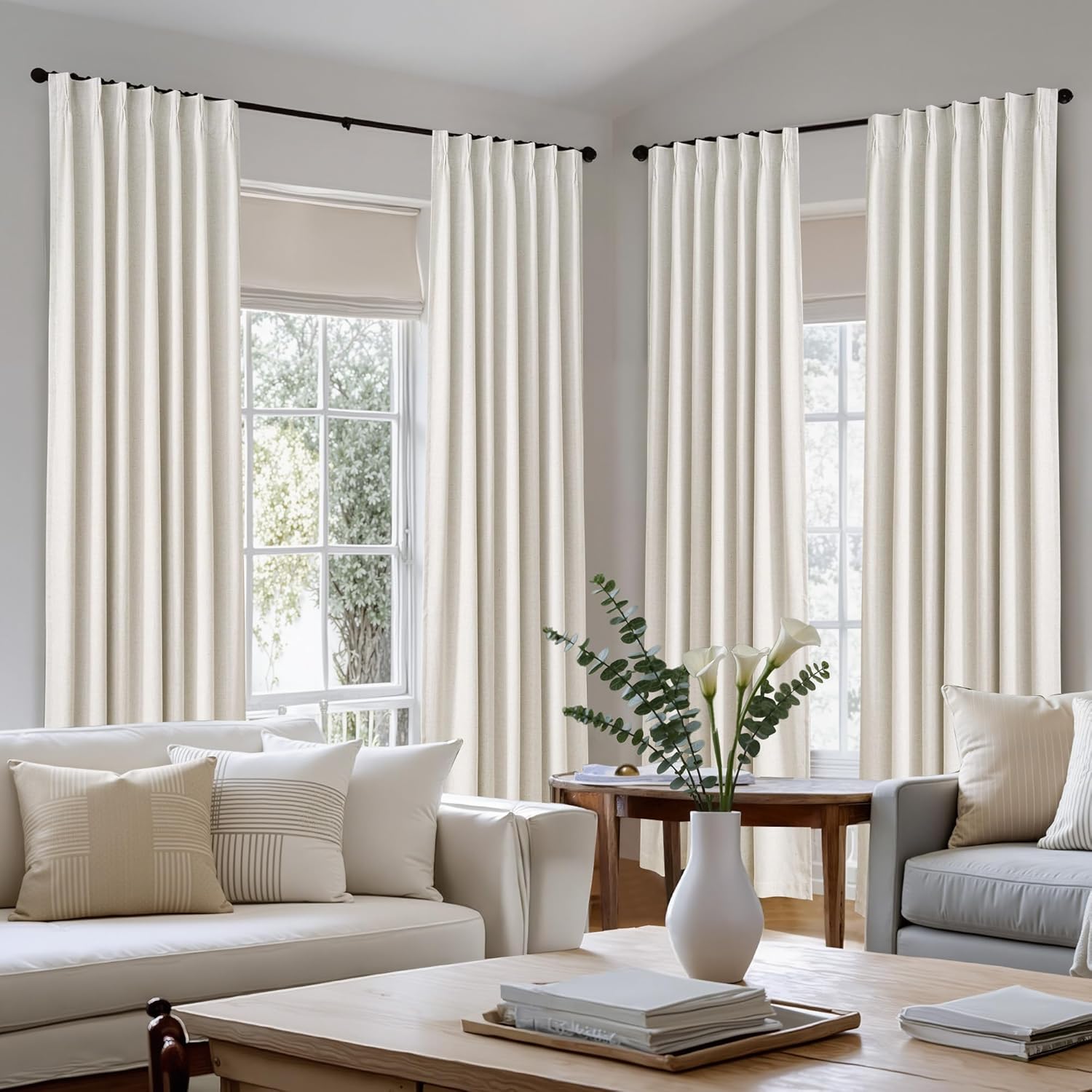Light beige linen pinch pleat curtains hanging on a black rod in a bright, modern living room.