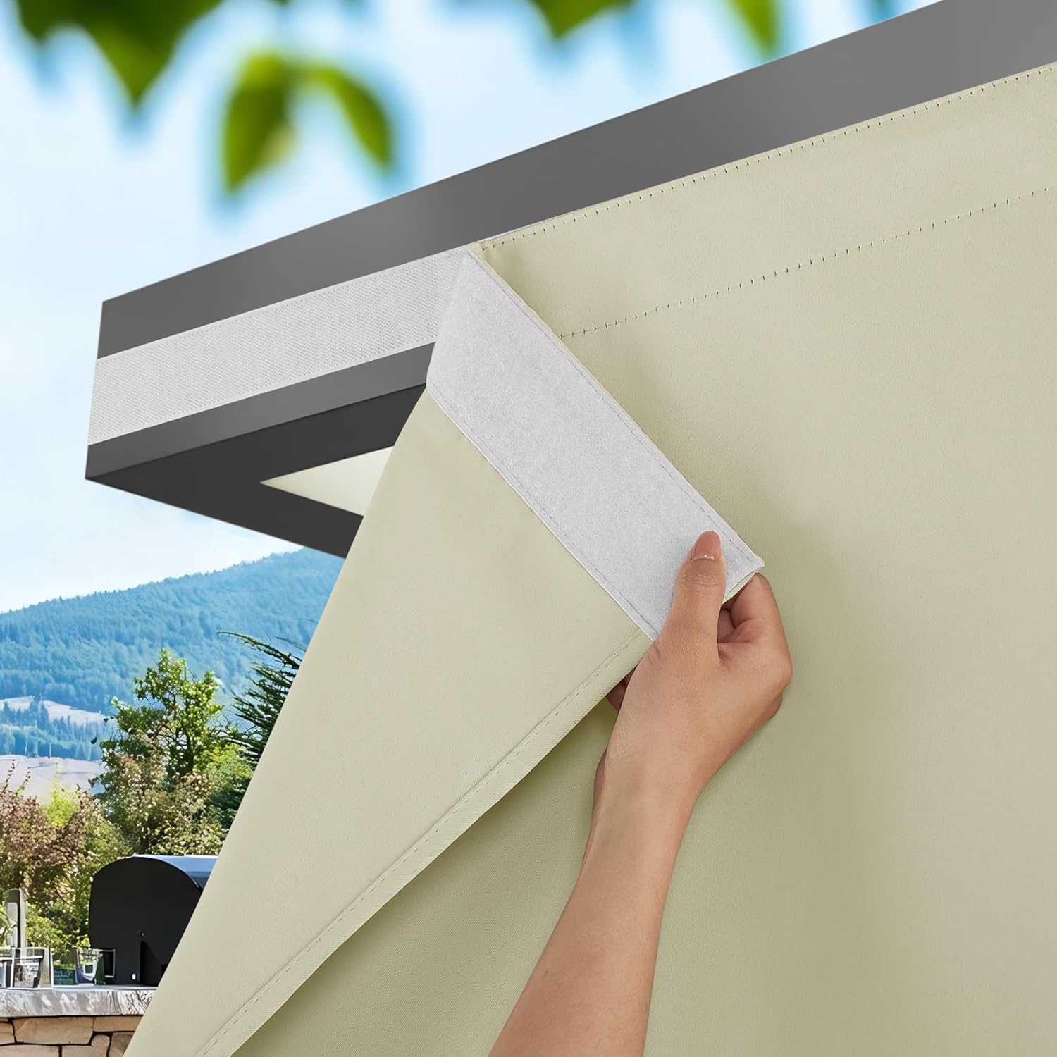 Hand attaching light beige outdoor shade panel to pergola with hook-and-loop fastener against sunny mountain view.
