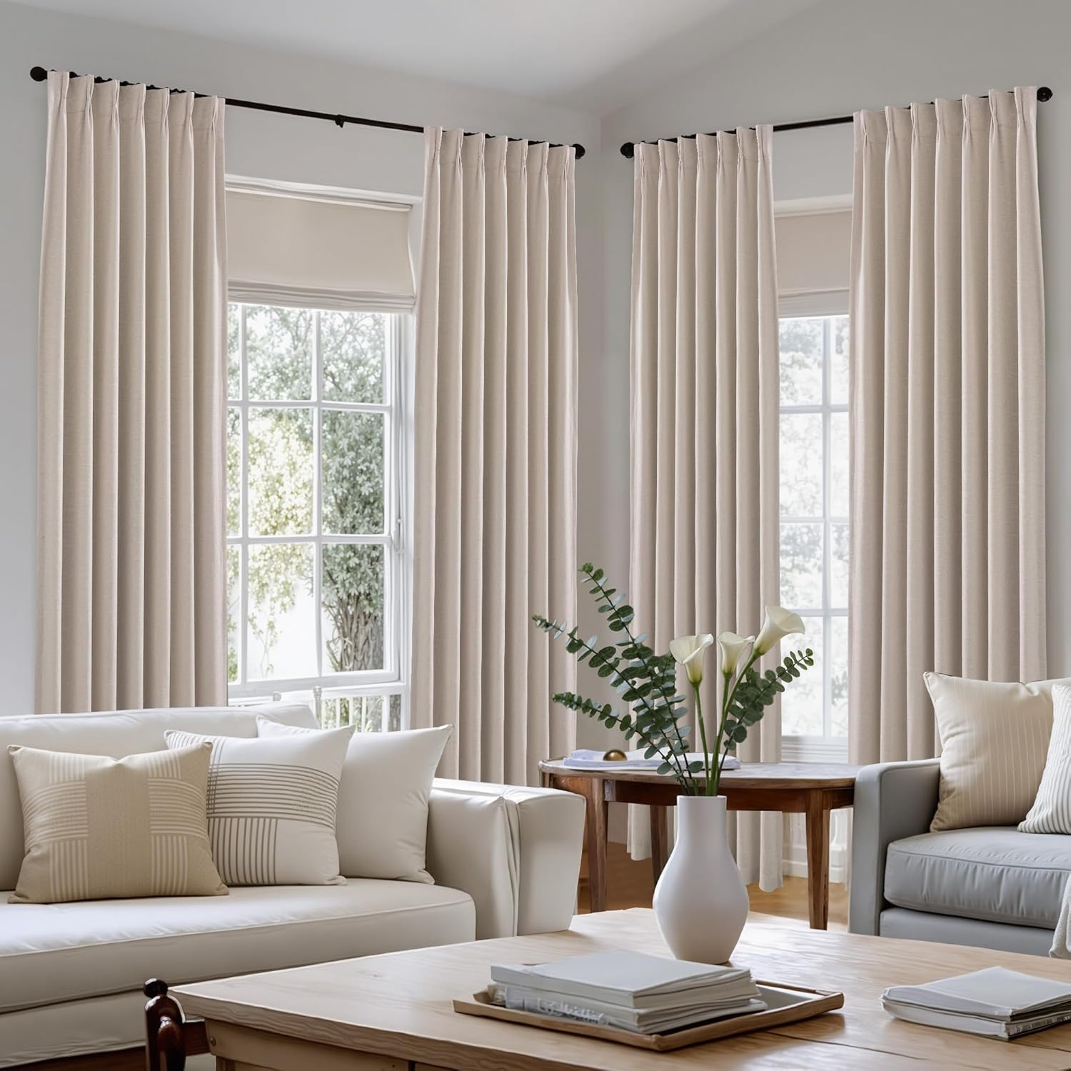 Elegant light beige pinch pleat drapes on black rods, framing tall windows in a bright, modern living room.